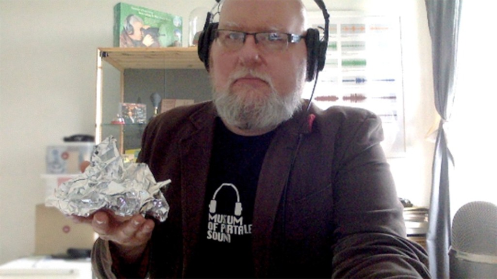 The MOPS Director, with a serious look on his face, wearing a pair of headphones while on a video call. In his right hand he holds what appears to be a crumpled up piece of aluminium foil which keenly resembles the preceding photographs of the Frank Gehry Commemorative Wing.